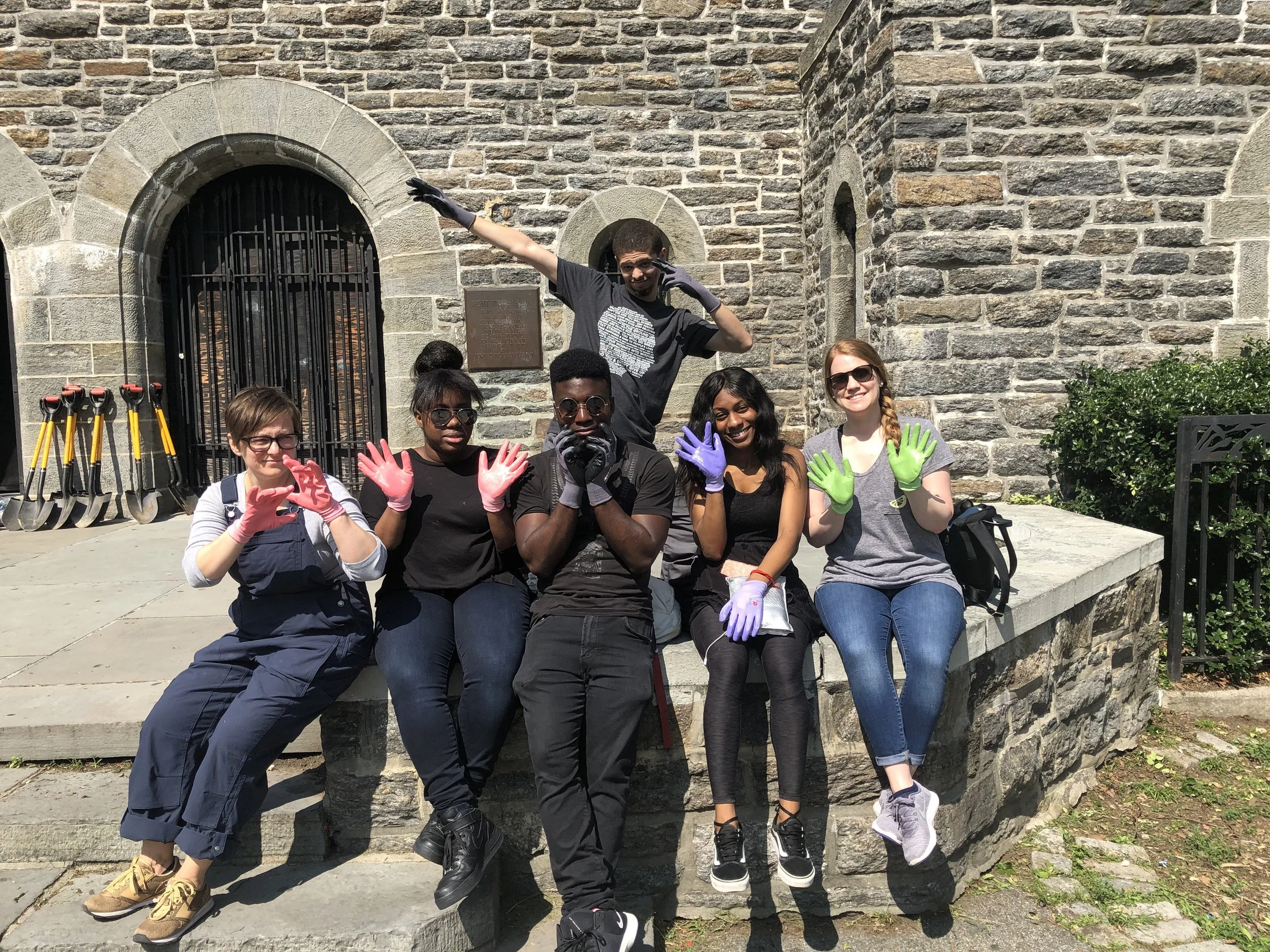 A group of seven diverse young people sitting on a stone wall, wearing colorful gloves, and posing for a photo in front of a historic stone building with an arched doorway. Some are smiling, making hand gestures, while one person stands behind them with sunglasses and gloves, striking a playful pose.