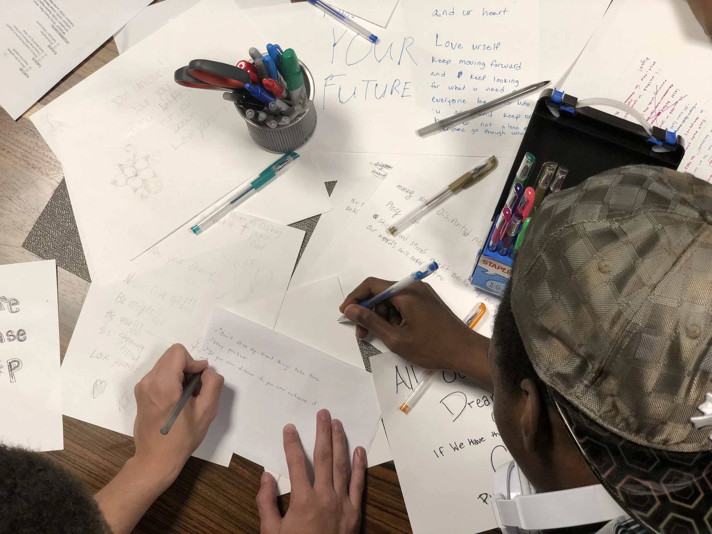 People gathering around a wooden table, writing and drawing on various sheets of paper with pens and markers, surrounded by stationery supplies.
