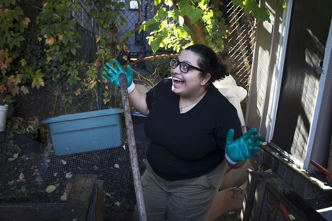 A woman with glasses and dark curly hair smiling, wearing gloves, standing in a garden with plants and a blue container, holding a gardening tool.