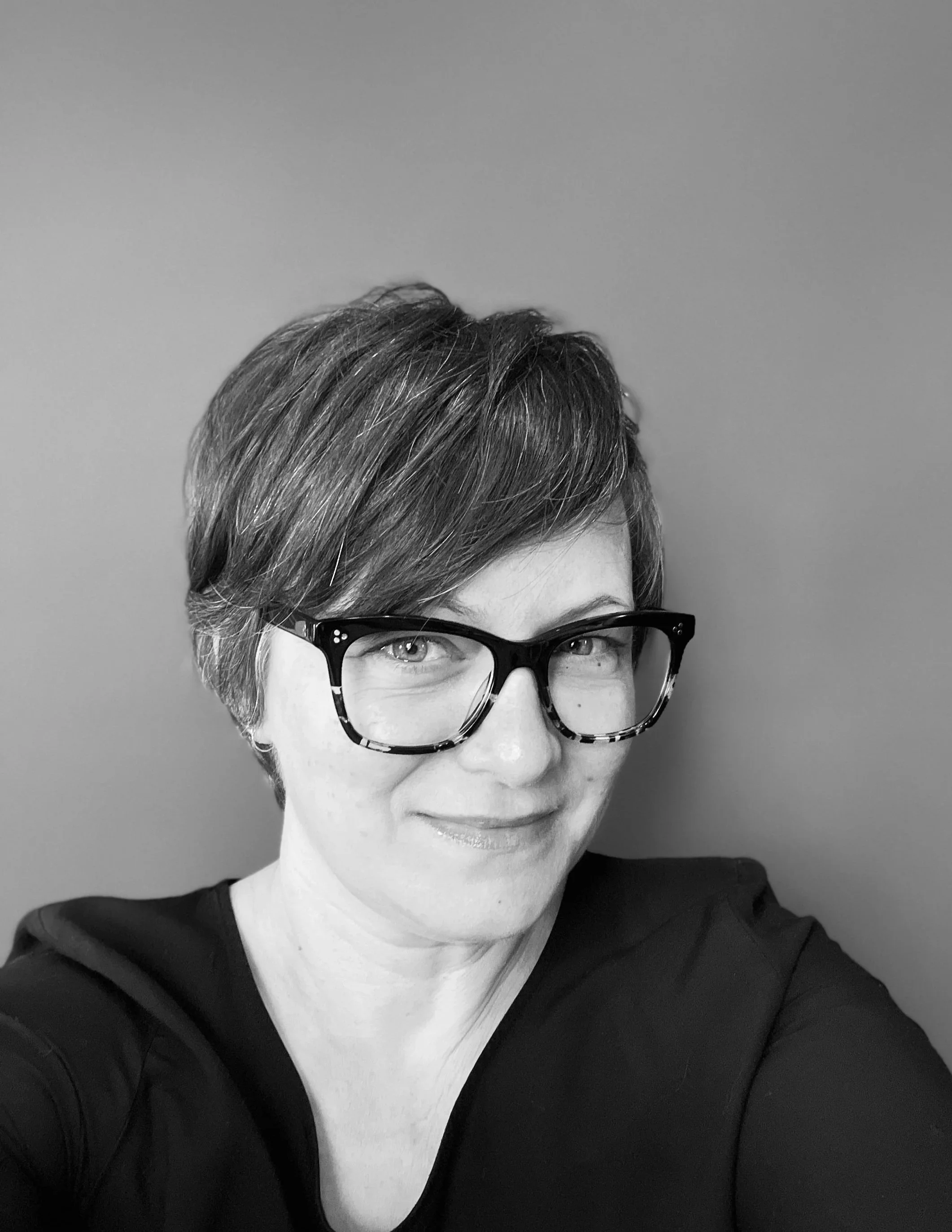 A smiling woman with shoulder-length hair wearing large glasses, indoors with a blurred bookshelf in the background.