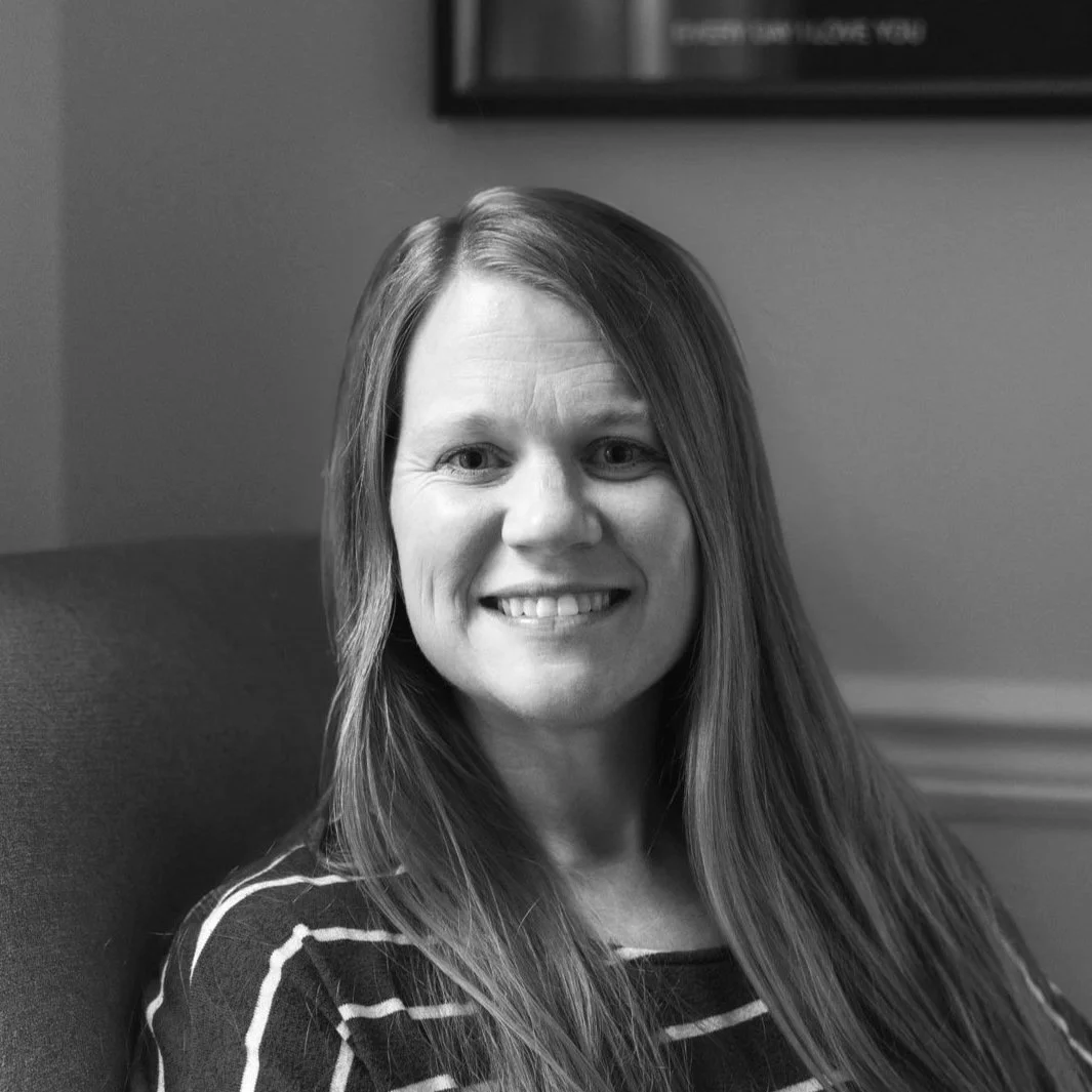 Black and white photograph of a smiling woman with long, straight hair, sitting in a chair indoors.