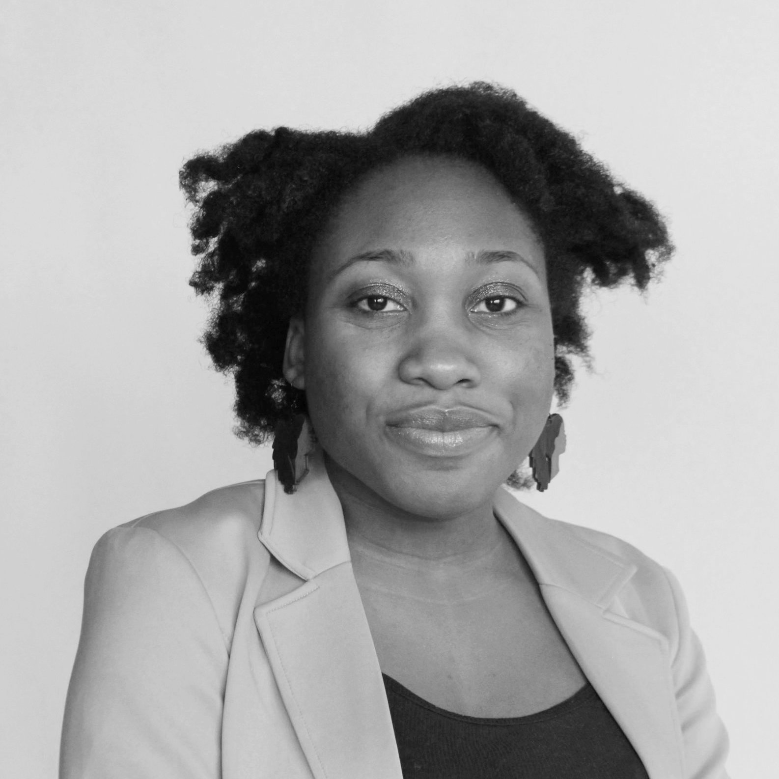 Black and white portrait of a woman with natural hair, wearing earrings and a blazer, looking at the camera with a slight smile.