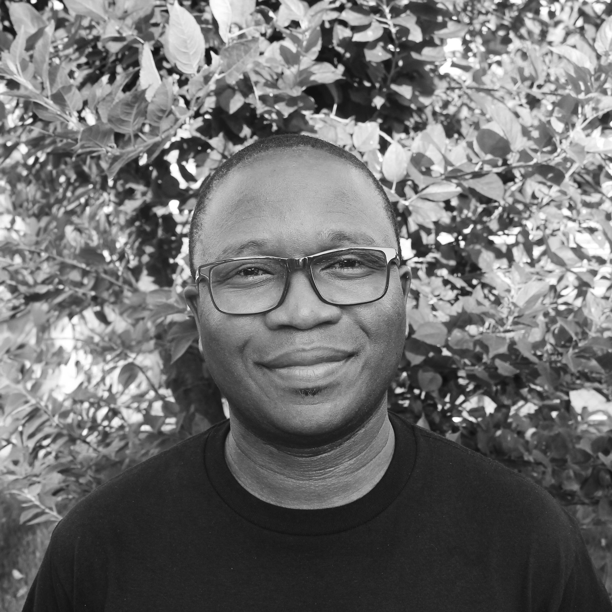 A man wearing glasses and a black shirt smiling outdoors with trees and bushes in the background.