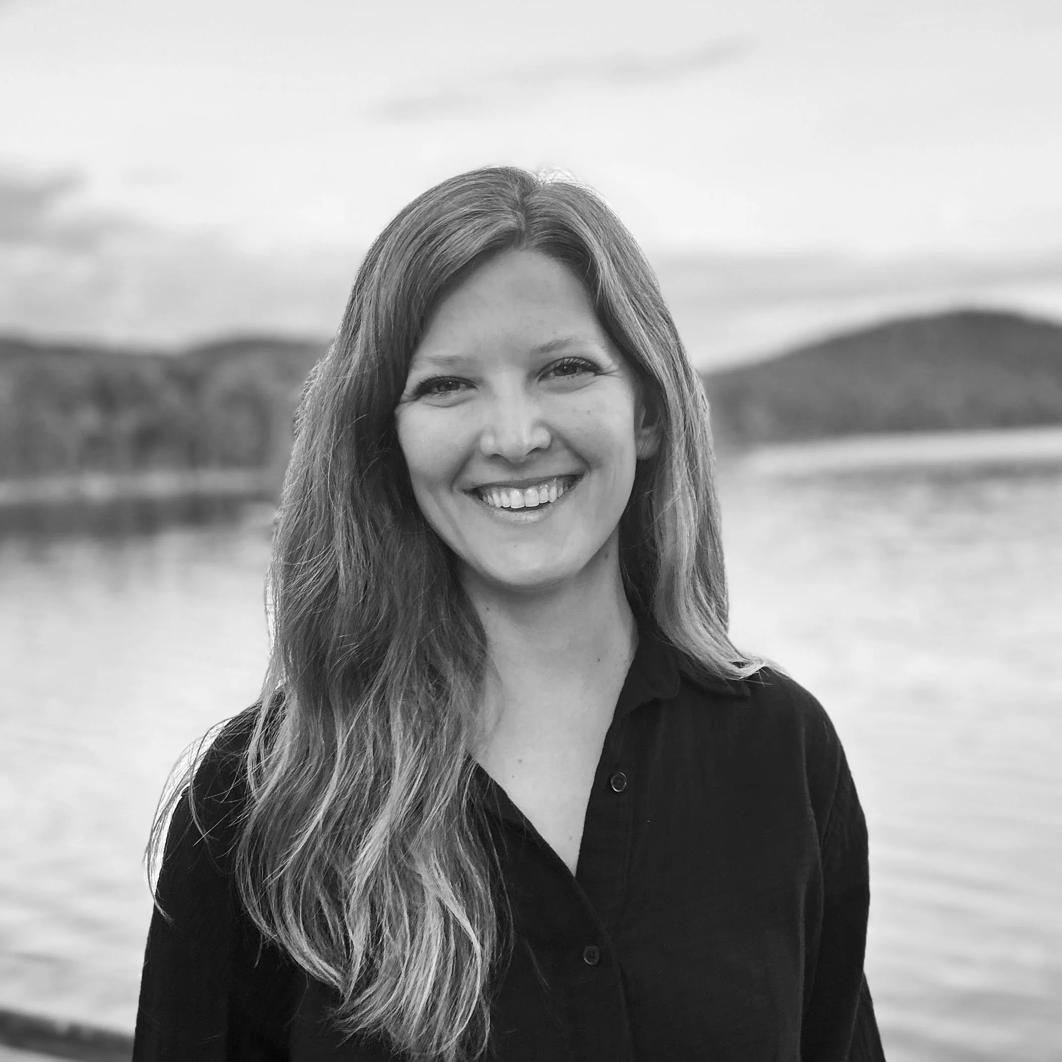 Black and white portrait of a smiling woman with long wavy hair, standing outdoors near a body of water with distant hills in the background.
