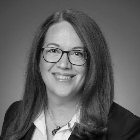 Black and white portrait of a smiling woman with long hair, glasses, and a blazer.
