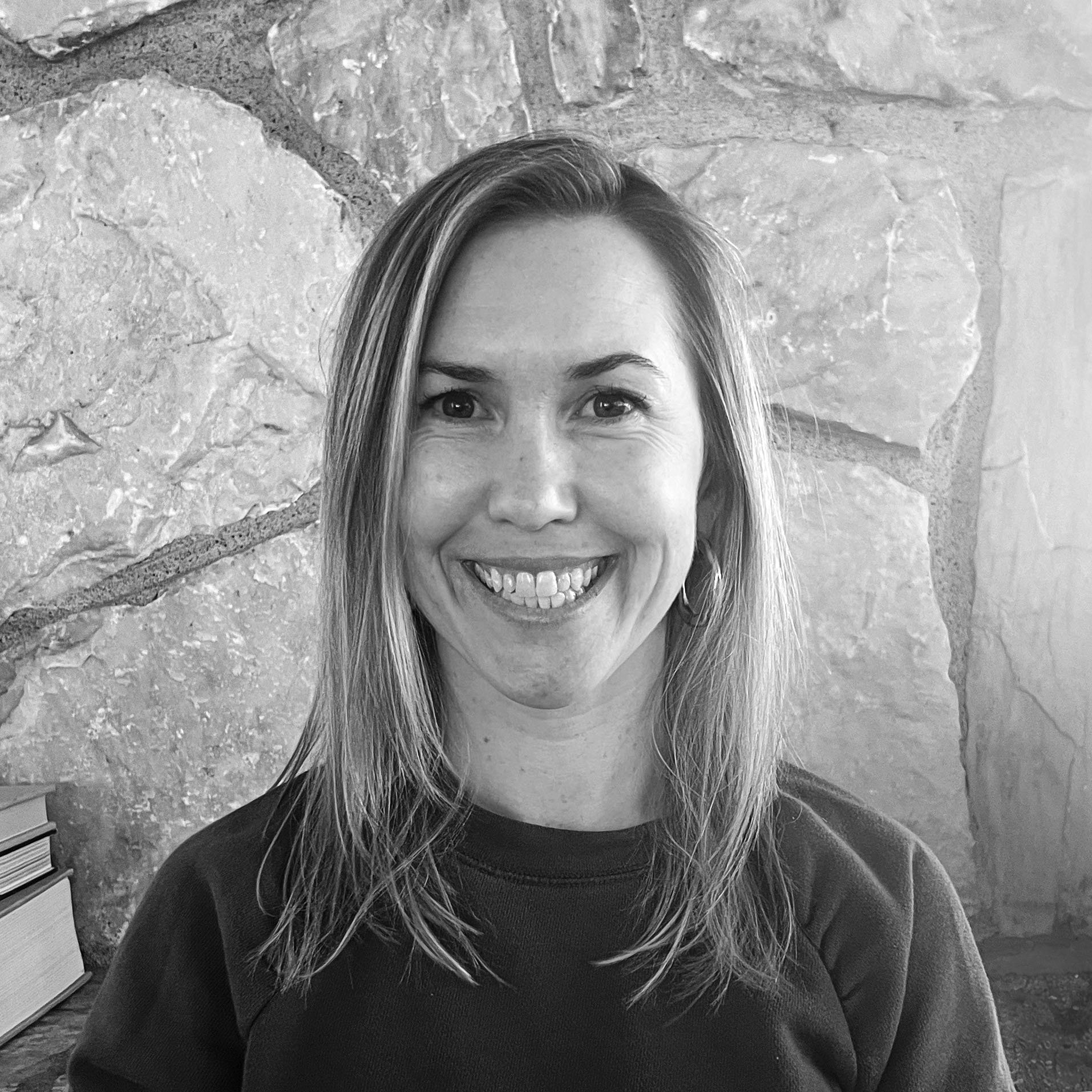 Smiling woman with shoulder-length hair in front of a stone wall, wearing a dark shirt.