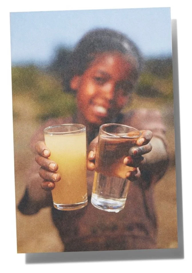 Young girl smiling outdoors holding two glasses of beverages, one yellowish and one clear.