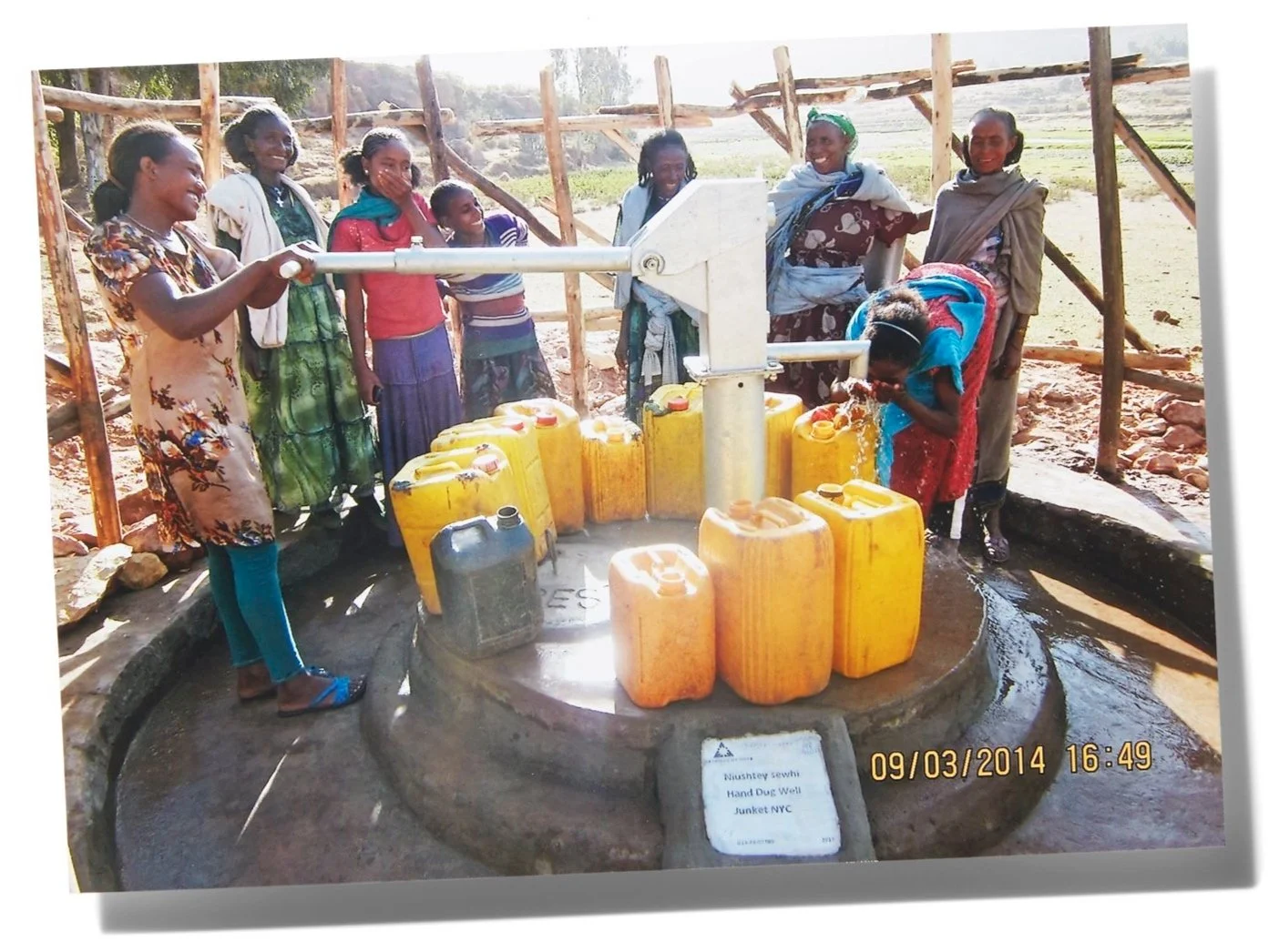 Group of women and girls gathered around a hand pump water well with yellow and black jerry cans, some women are filling water, others are smiling and observing, in a rural outdoor setting.