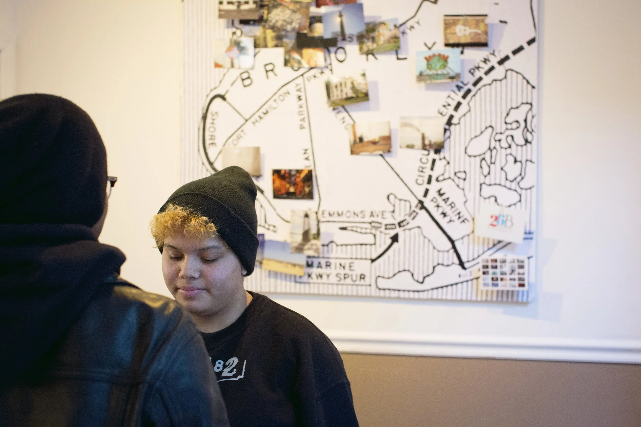 Two people standing in front of a large map with photos and labels on a wall, one with a black hoodie and the other with a black beanie and short curly blonde hair.