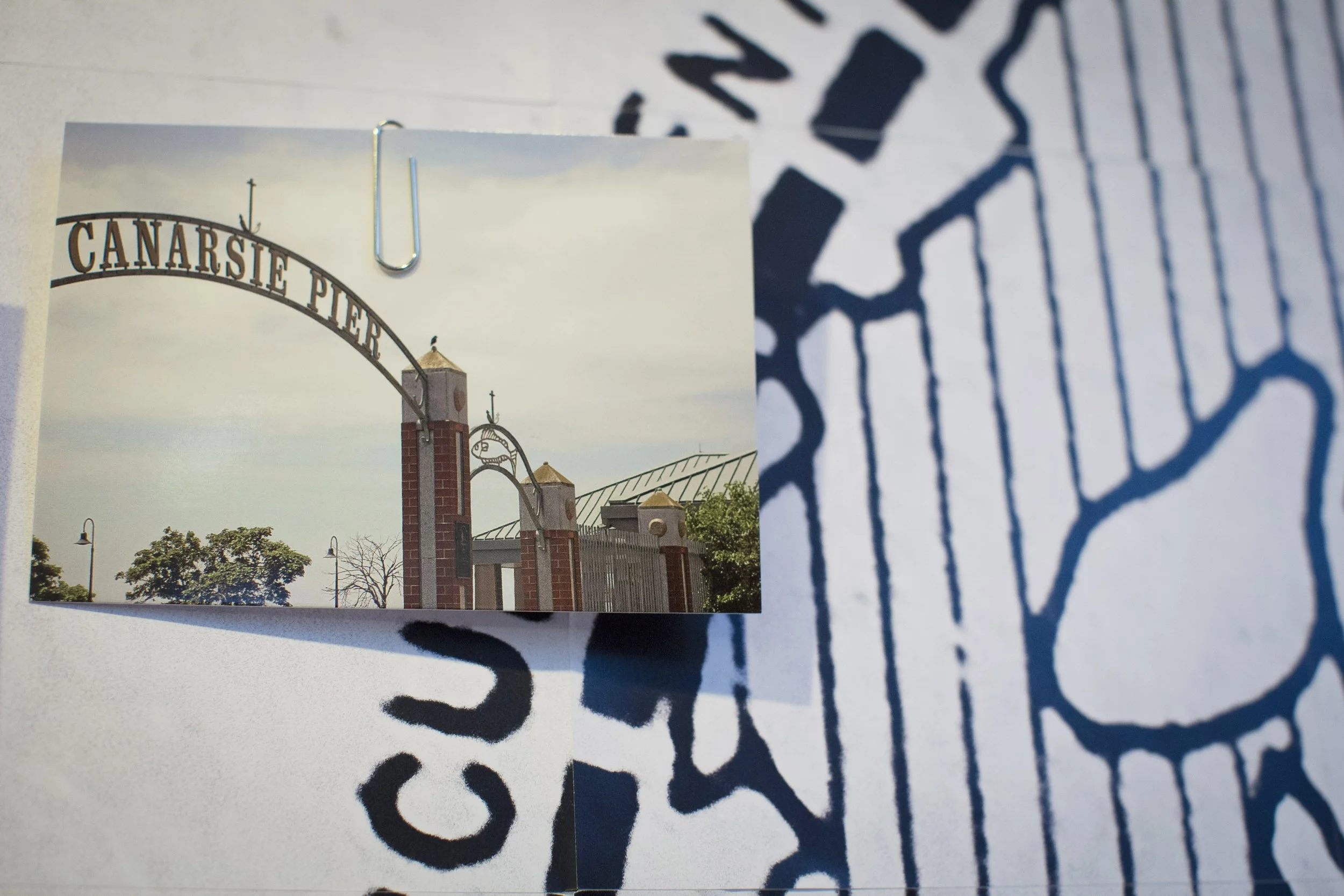 A photograph of the entrance to Canarsie Pier in Brooklyn, New York, showing a large metal arch sign with the words 'CANARSIE PIER' and brick columns underneath, with trees and a cloudy sky in the background.