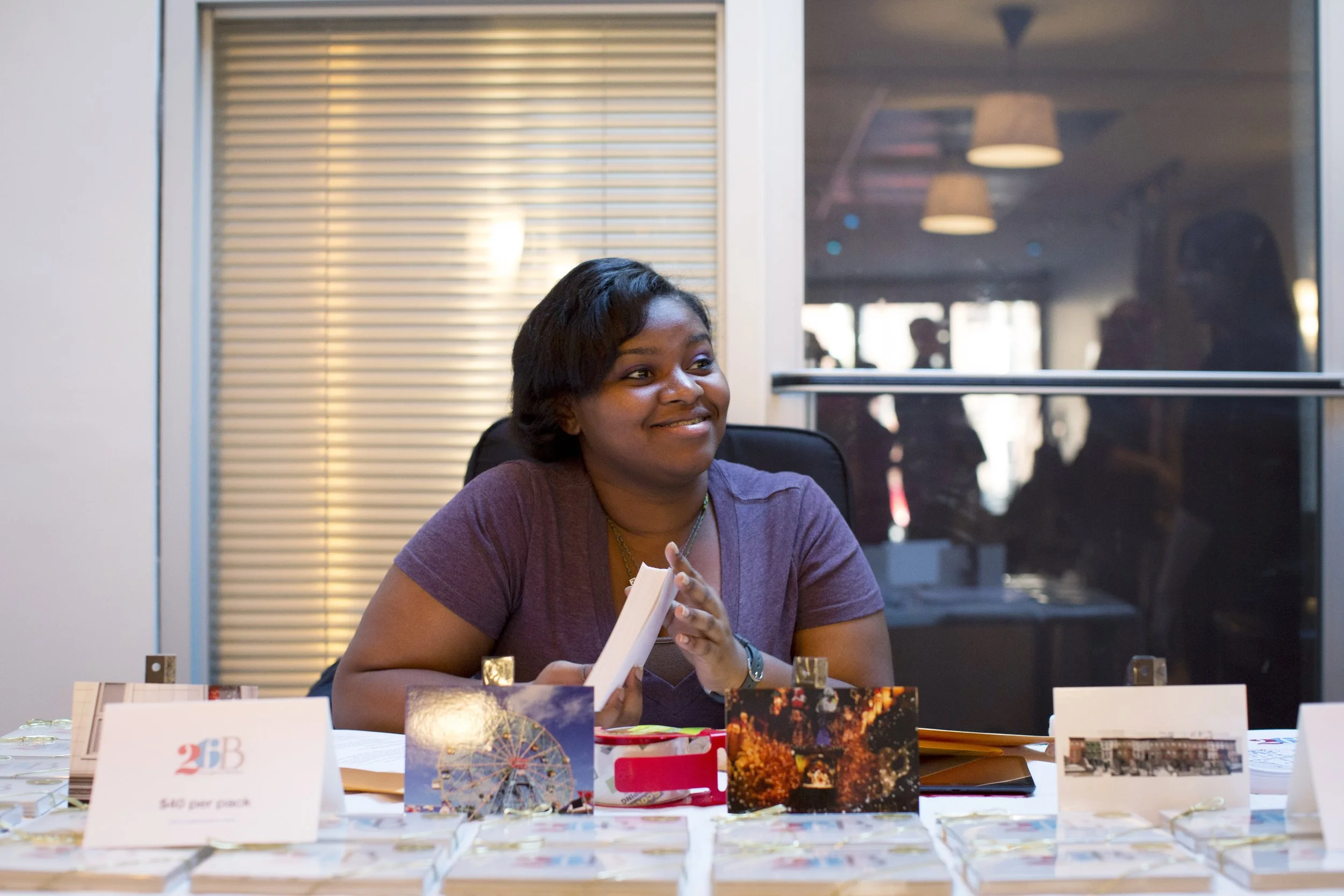 A woman smiling and sitting at a table with postcards and paper items, holding a small booklet, in a room with a window behind her and blurred figures in the background.
