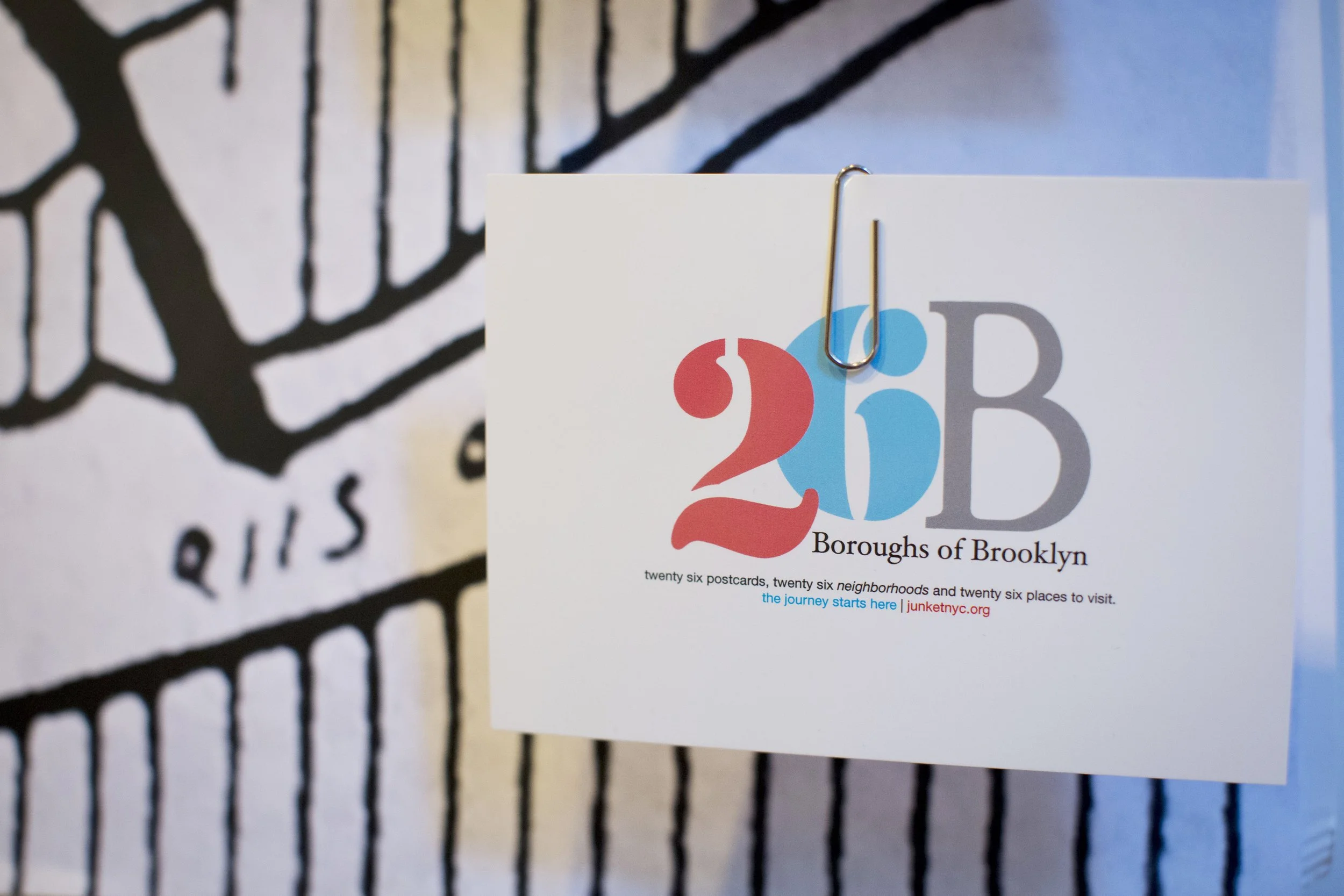 A flyer promoting Brooklyn neighborhoods, with the title '26 Boroughs of Brooklyn' and details about postcards and places to visit, attached with a metal paperclip. In the background, a black and white drawing of a subway map is partially visible.