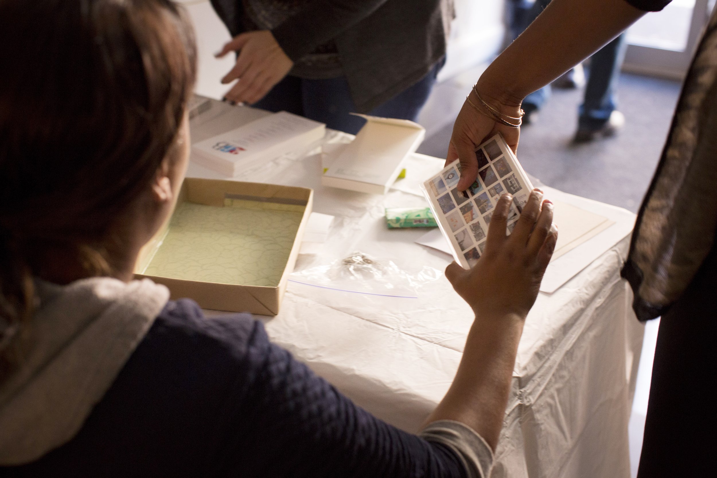 A person with reddish-brown hair and a gray hoodie looks at a collection of small photographs or cards with another person in front who is handing over some cards at a table with a white cloth cover.