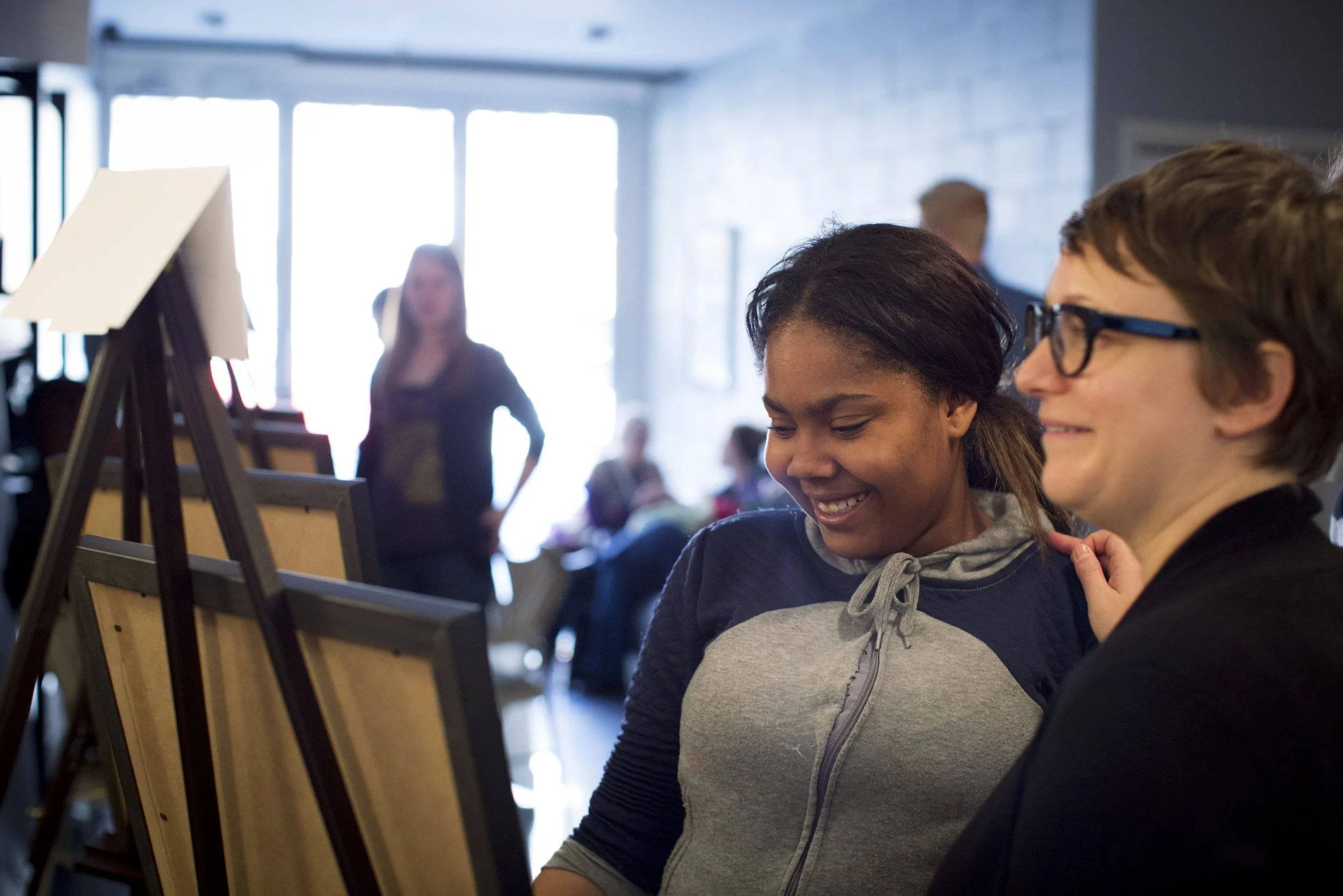 Two women smiling and enjoying each other's company at an art gallery or studio, with several empty picture frames and a woman in the background near a large window.