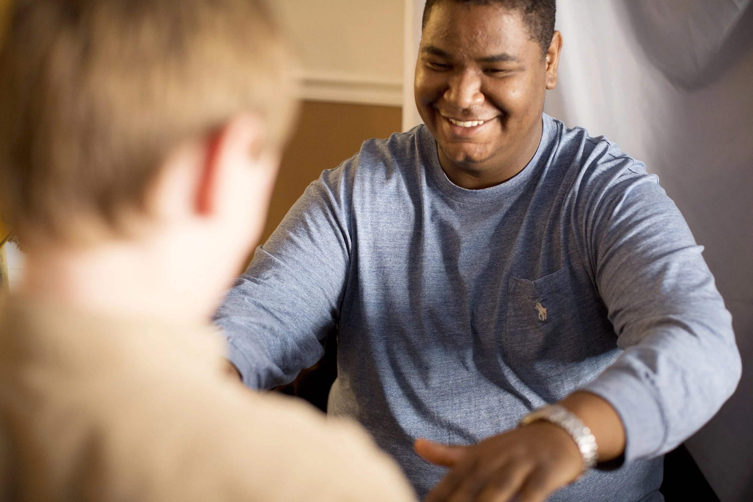 A smiling man in a blue sweater engaging in a conversation with a woman whose face is out of focus in the foreground.