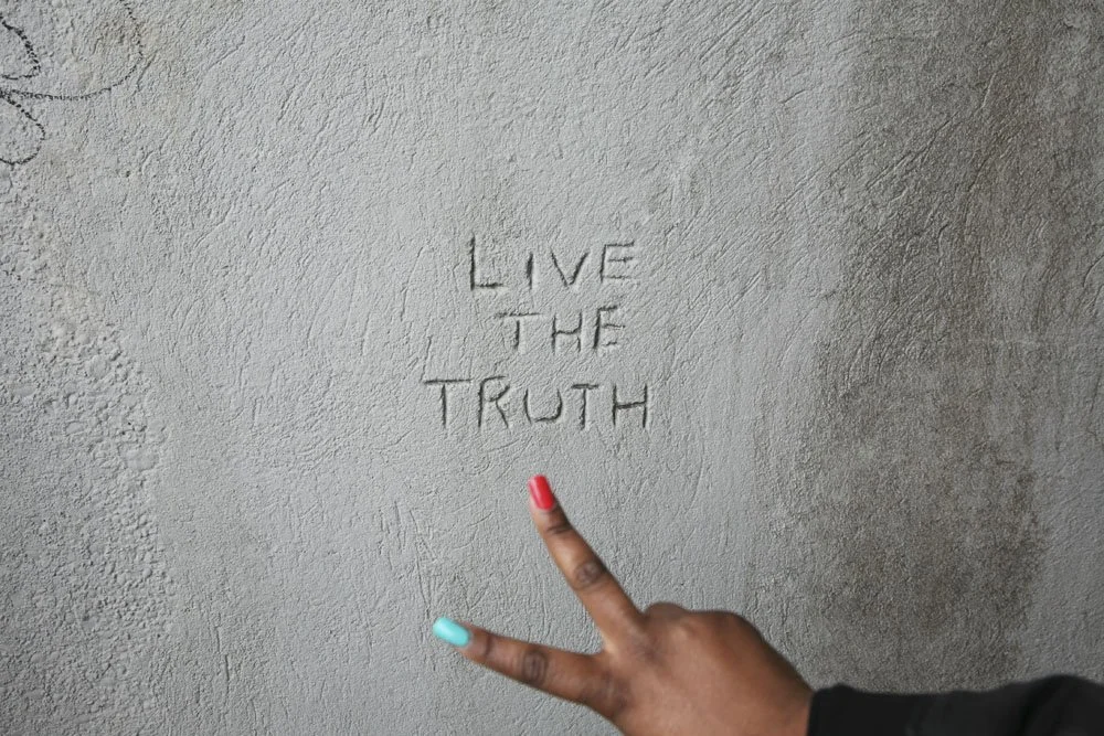 A wall with the phrase "Live the Truth" carved into it and a person's hand making a peace sign pointing towards the phrase.