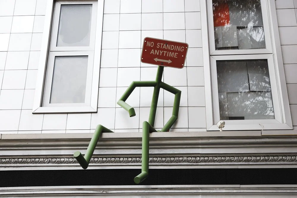 A green sculpture of a seated figure with its legs crossed, positioned on a building's ledge, with a red sign that reads 'No Standing Anytime' attached as the figure's head.