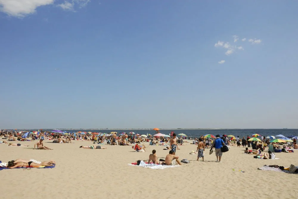 Crowded beach with many people sunbathing, walking, and using umbrellas under a clear blue sky.