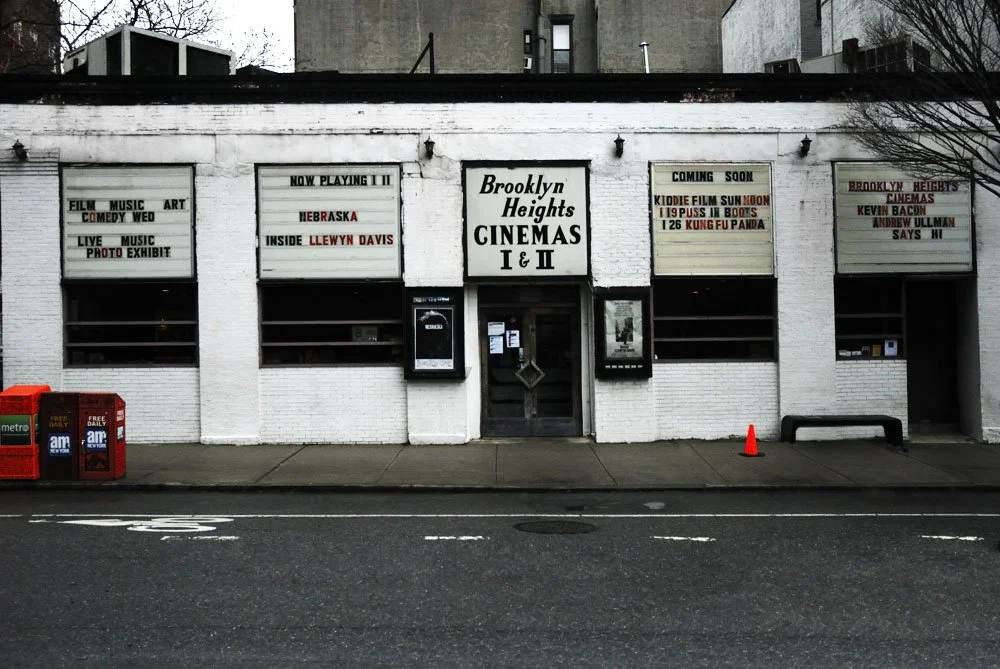 White brick building with signs indicating Brooklyn Heights Cinemas I & II, upcoming movies, and events, with newspaper boxes and a parking cone on the sidewalk.