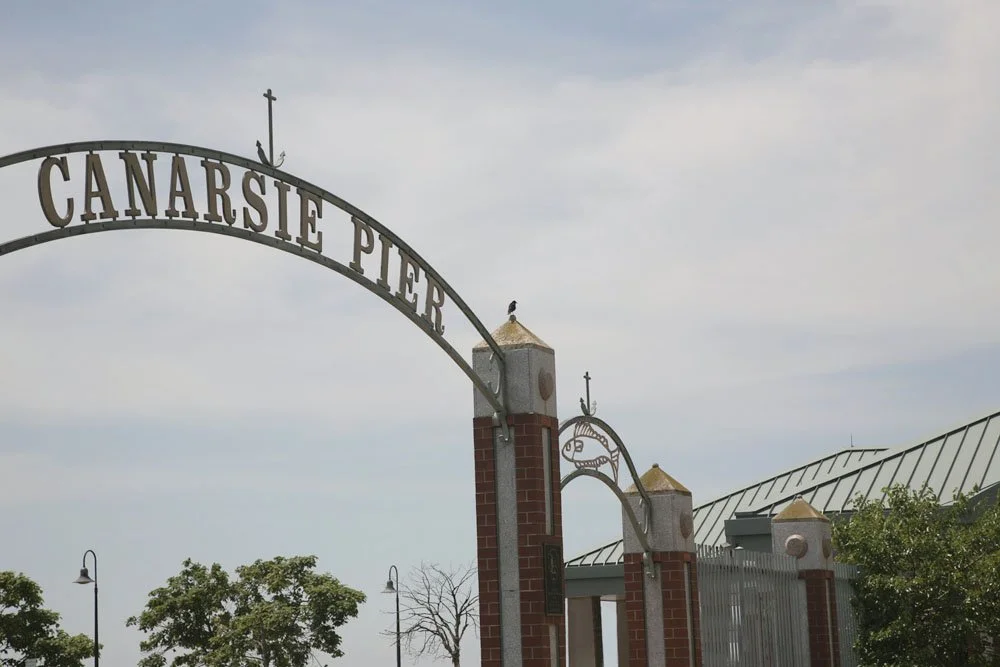 Archway with the words 'Canarsie Pier' over brick and metal columns, with trees and a cloudy sky in the background.
