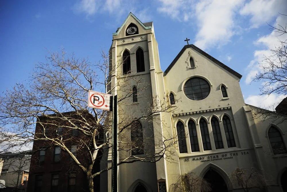 A white Roman Catholic church named St. Mary Star of the Sea with a tall clock tower, surrounded by leafless trees and a no-parking sign in the foreground, under a partly cloudy sky.