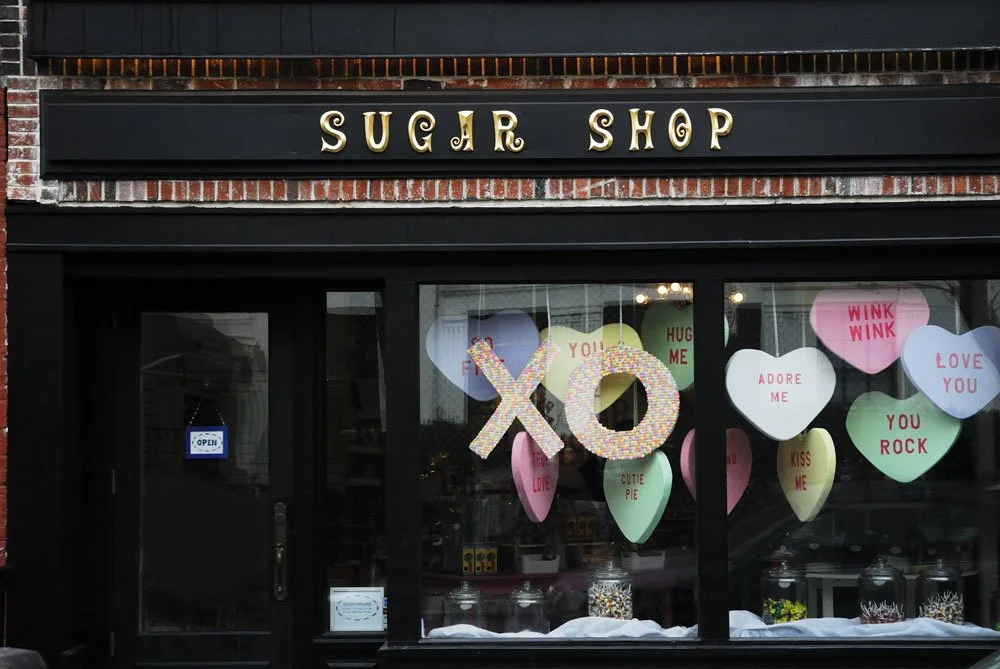Front of a sugar shop with a black sign that reads "SUGAR SHOP" in gold letters. The window is decorated with large paper hearts with phrases like "WINK WINK," "LOVE YOU," "ADOR ME," and "YOU ROCK." There are two large glittered 