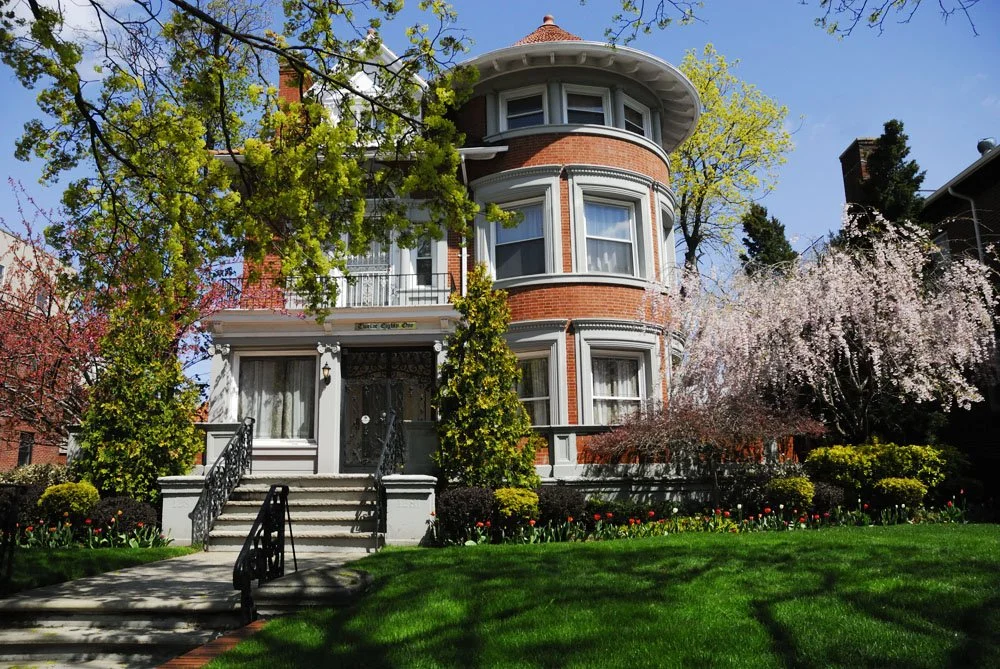 A large, historic red brick house with a rounded turret, surrounded by blooming trees, lush green lawn, and flowering bushes, under a clear blue sky.