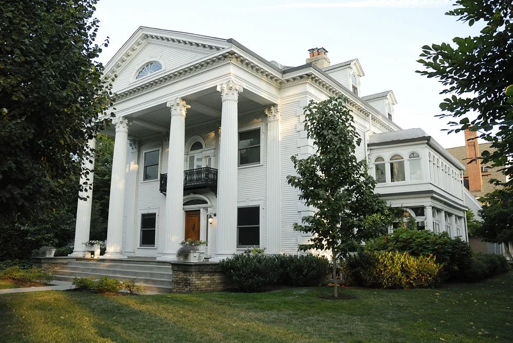 A large white historic house with tall columns and a porch, surrounded by trees and a green lawn.