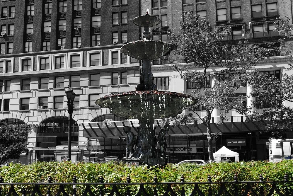 A black and white urban scene featuring a fountain with water cascading from multiple tiers, surrounded by greenery and located in front of a large building with multiple stories and numerous windows.