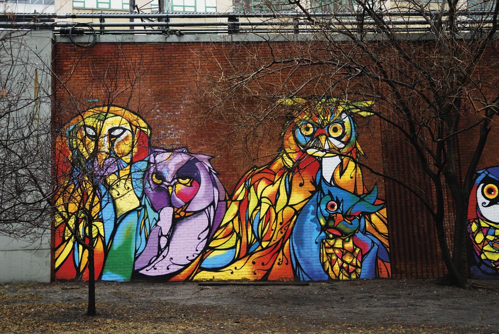 Colorful graffiti mural of various stylized animal faces painted on a brick wall, including an owl, a lion, a cat, and a fox, with leafless trees in front.