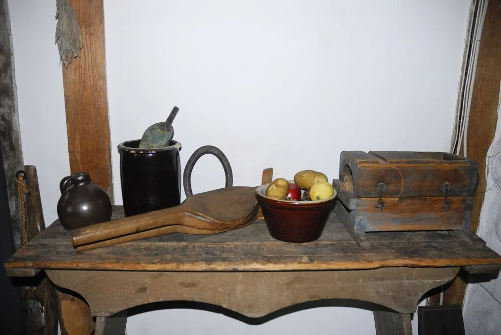 Rustic wooden table with vintage household items including a black jug, a wooden rolling pin, a large paddle, a bowl of apples and lemons, and a wooden chest. The background features a white wall with a wooden beam.