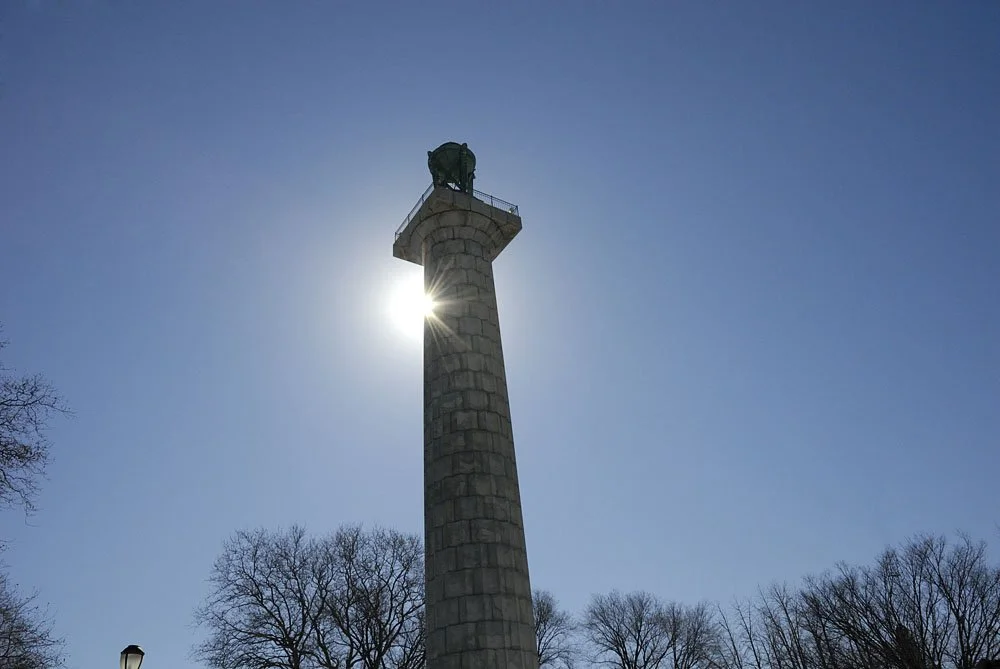 A tall stone monument with a small platform at the top, with the sun partially visible behind it and bare trees around the base under a clear blue sky.