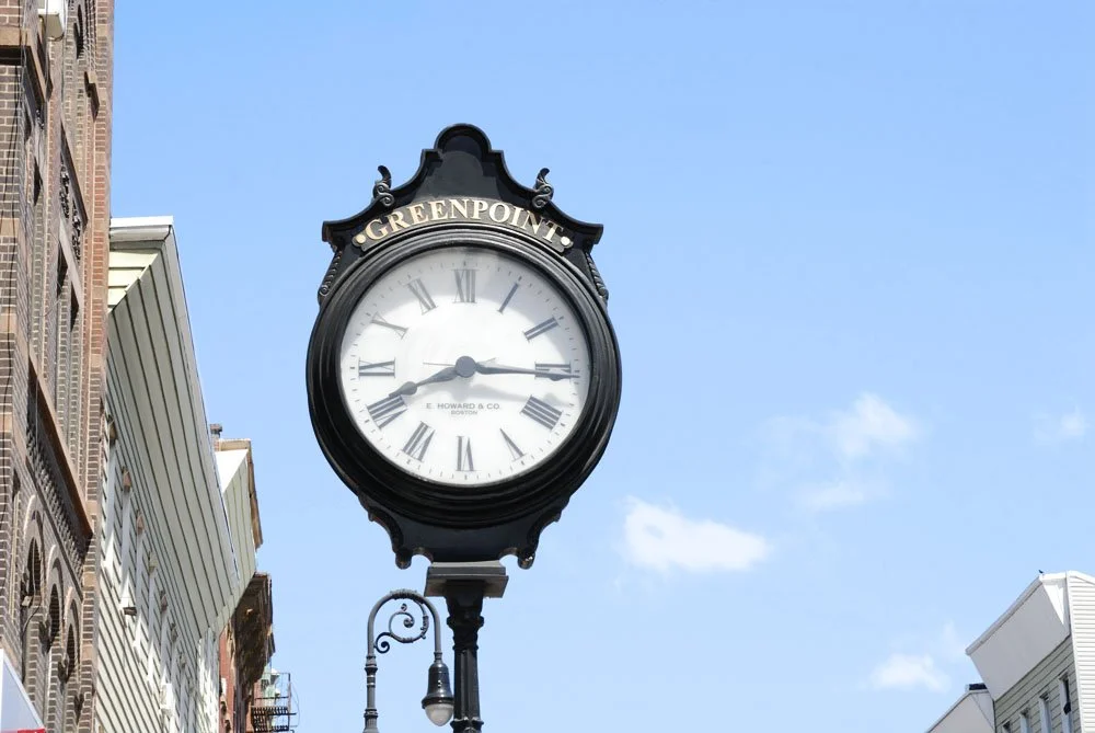 Street clock with the words 'Greenpoint' at the top, showing the time at approximately 3:17, against a bright blue sky with some clouds, surrounded by buildings.