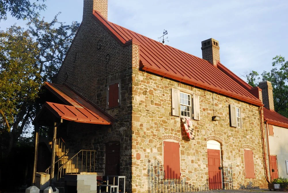 A historic stone house with a red metal roof, wooden shutters, and brick chimney, surrounded by trees under a clear sky.