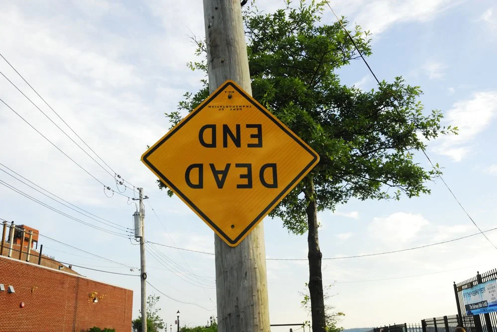 A yellow diamond-shaped road sign reading 'DEAD END' is mounted upside-down on a wooden utility pole, with power lines, a brick building, a green tree, and a partly cloudy sky in the background.