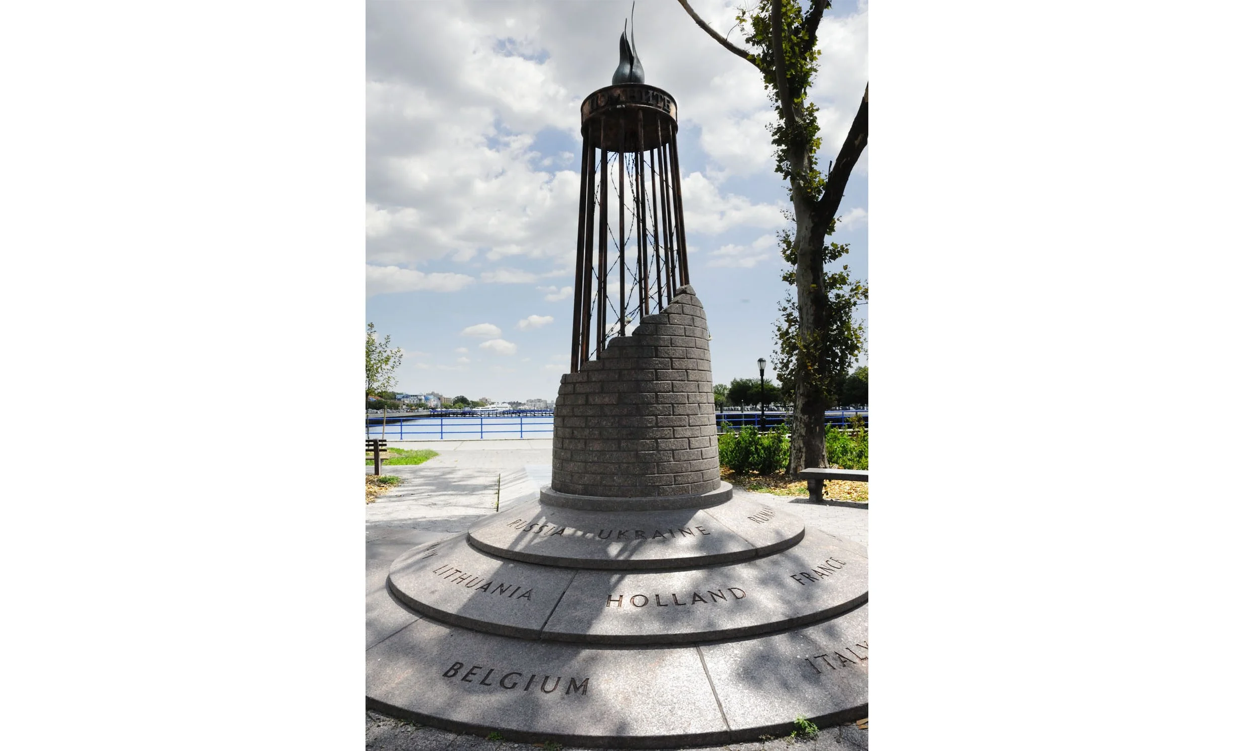 A memorial monument with a brick base and metal top, surrounded by a circular stone platform inscribed with country names like Belgium, Ukraine, and France, located outdoors near a river with trees and benches around.