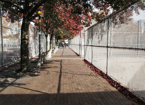 A paved sidewalk lined with trees on the left and a chain-link fence on the right, with a few people walking in the distance during daytime.