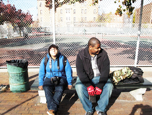 A man and a young girl sitting on a park bench near a chain-link fence, with a baseball field in the background. The girl is wearing glasses and a blue jacket with a backpack, while the man is wearing a black jacket and red gloves, looking down.
