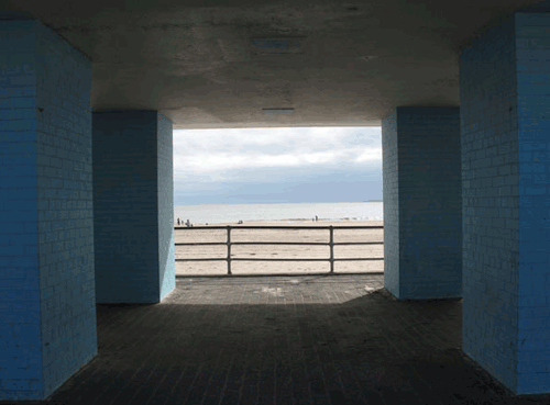 View of a beach seen through an underpass with blue brick columns and a railing, overcast sky.