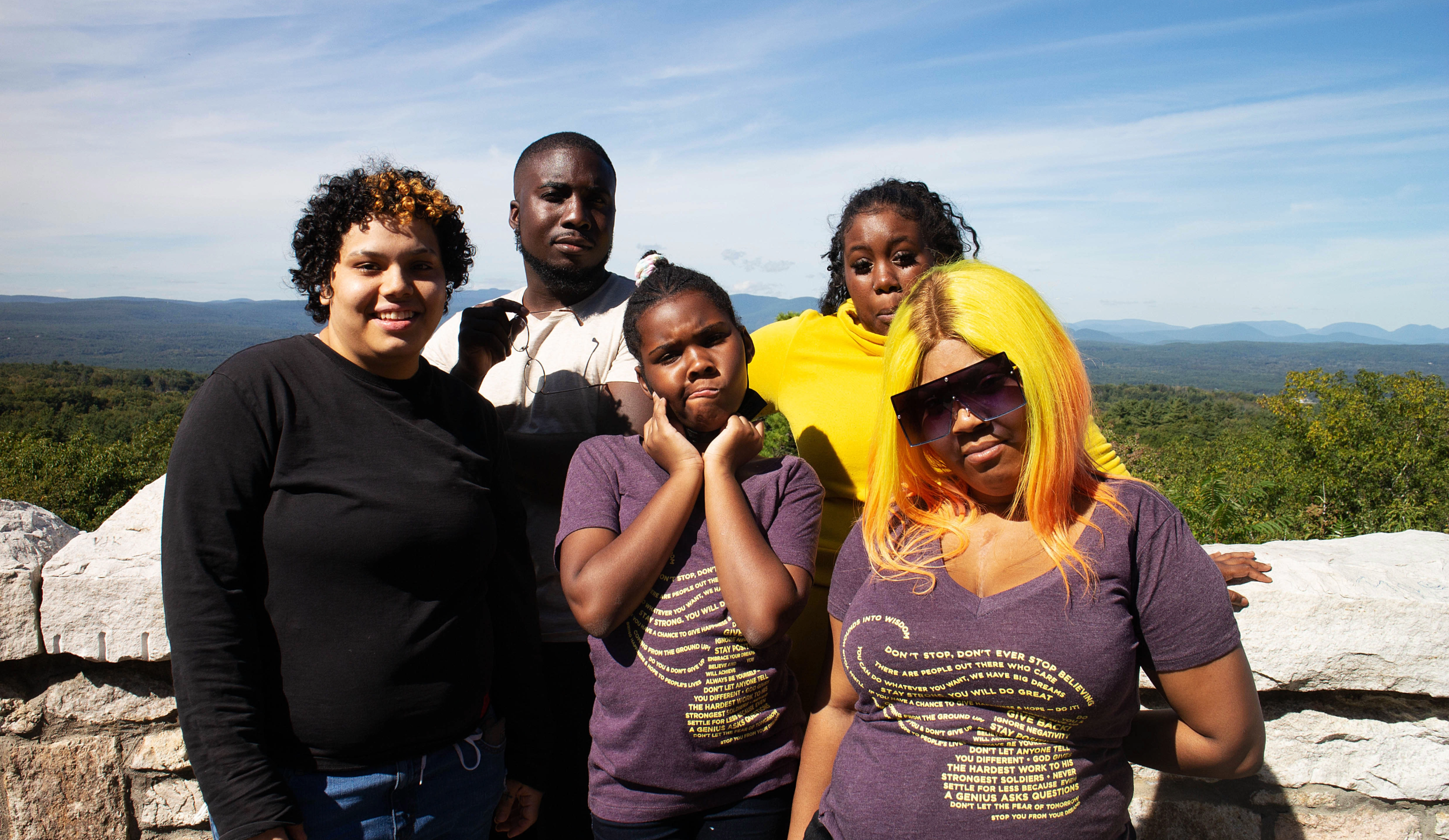 Group of six diverse young women and girls outdoors on a clear day, standing near a stone wall with a scenic mountain and forest background.
