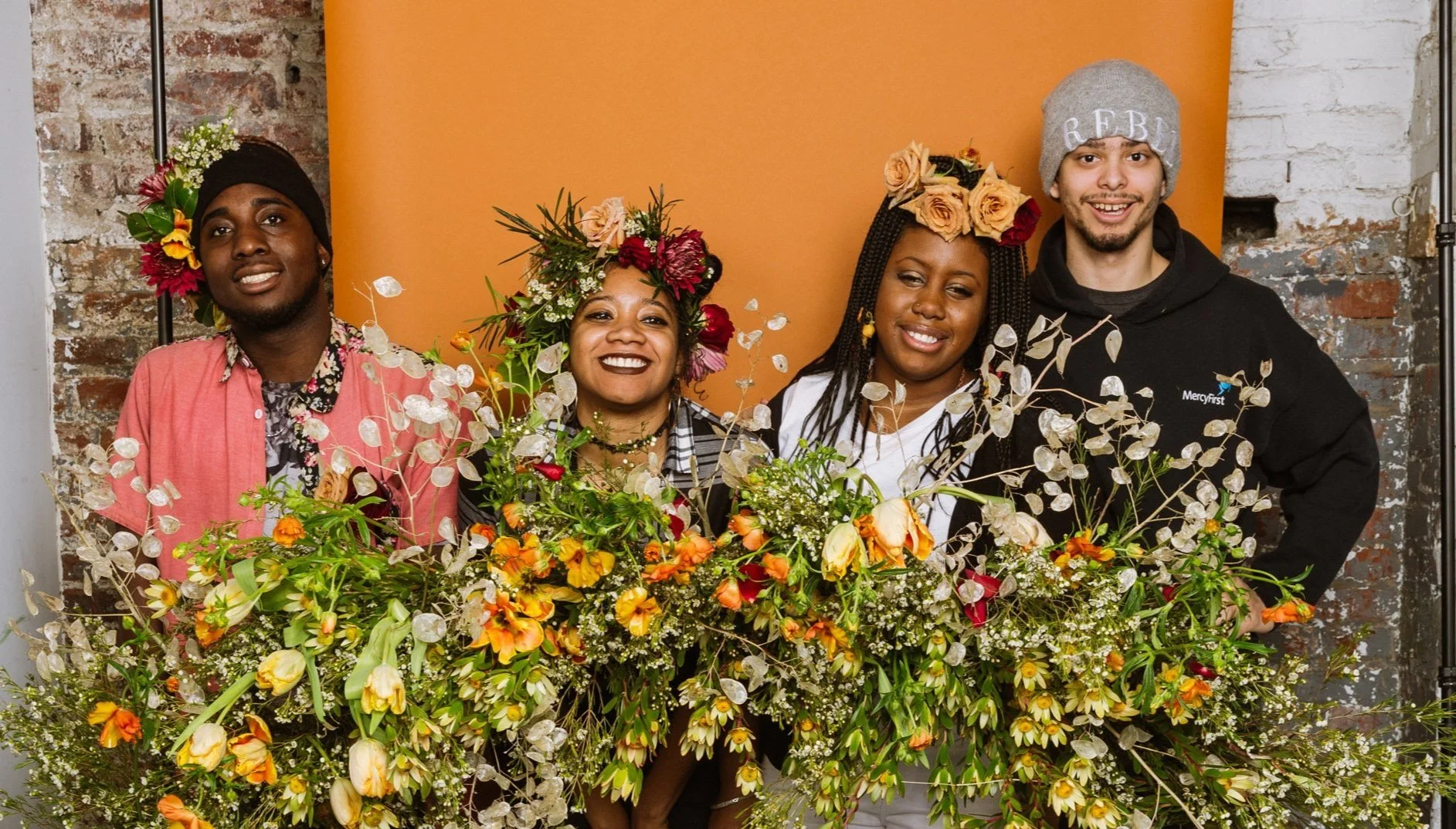 Four people standing behind a large floral arrangement, smiling, with a brick wall and orange backdrop behind them.