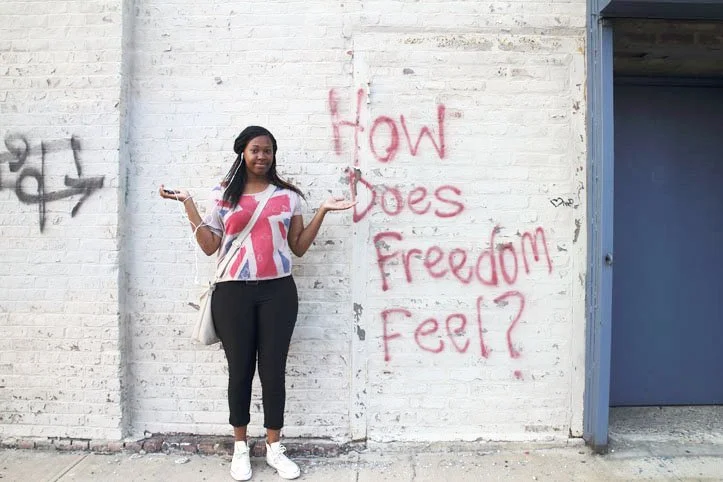 A young woman standing in front of a white brick wall with graffiti that says 'How does Freedom Feel?' She is wearing a T-shirt with the Union Jack, black pants, and white sneakers, with her arms out to the sides and a neutral expression.