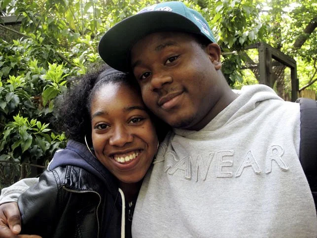 A young couple smiling, standing close together outdoors with green foliage in the background.