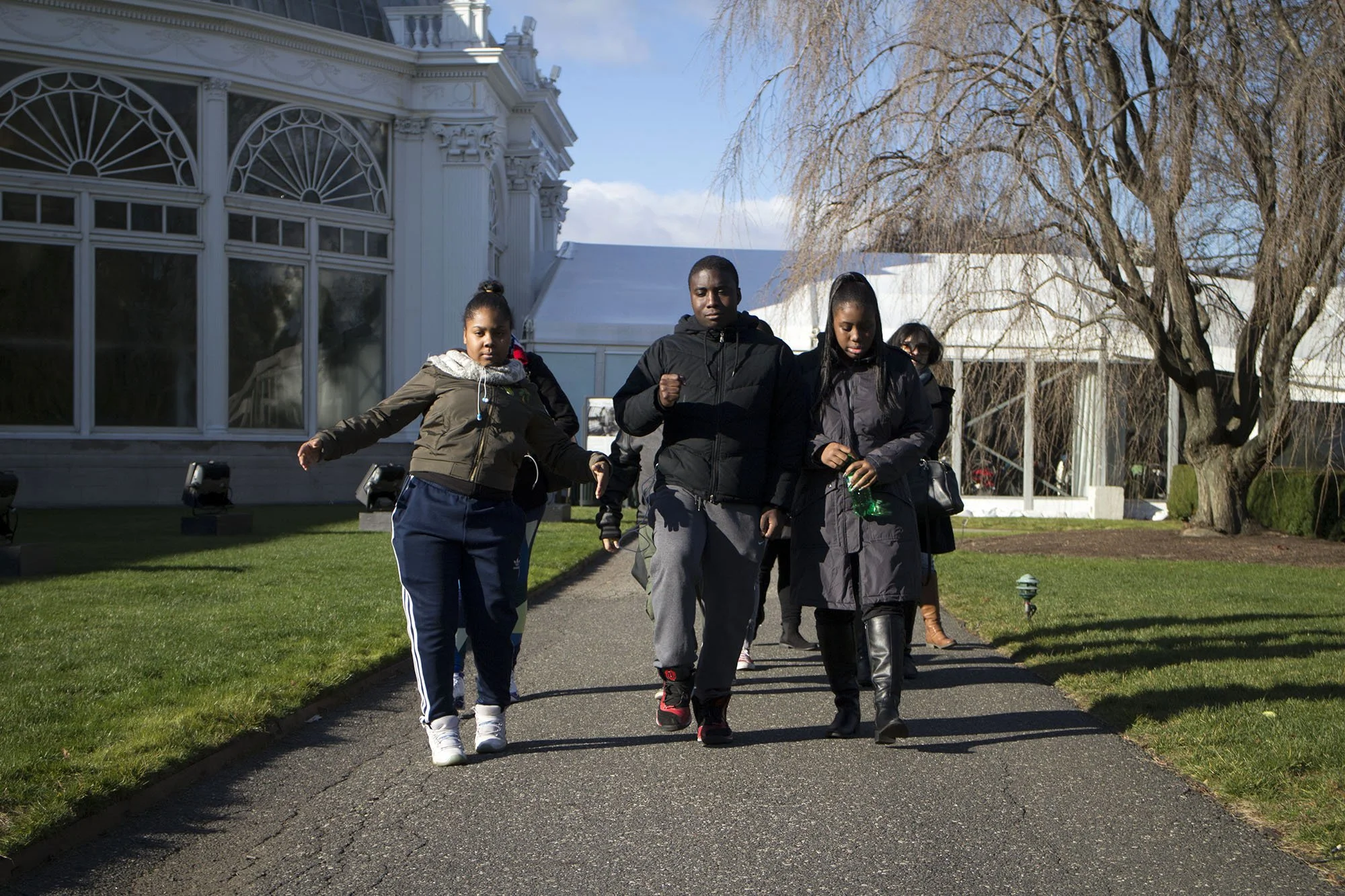 A group of young people walking on a paved path outdoors on a sunny day, with a white building, leafless trees, and a green lawn in the background.