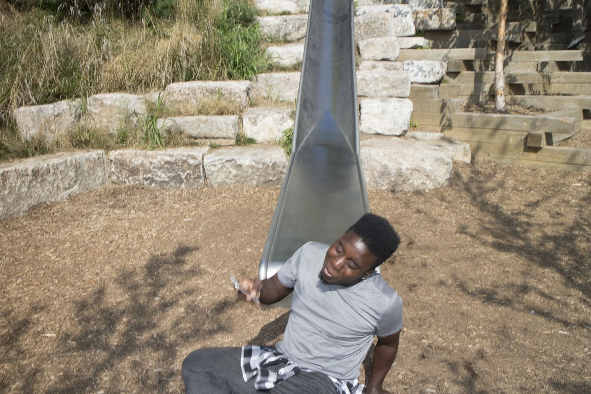 Young man in gray shirt and black shorts sitting on ground near a slide in a playground area with stone and wooden steps.