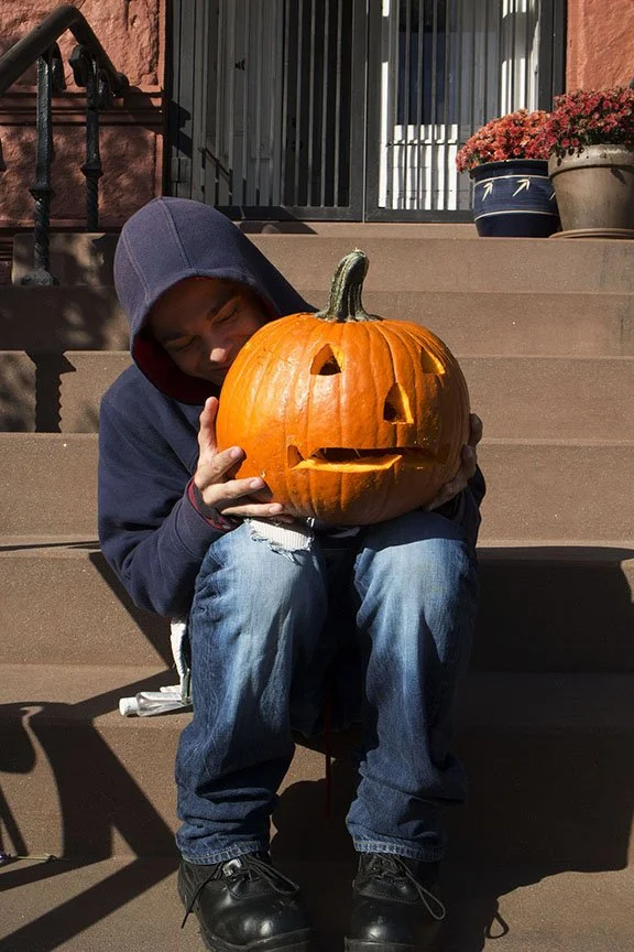 Young boy sitting on outdoor stairs, holding a carved pumpkin with a happy face, wearing a hooded sweatshirt and jeans, enjoying a fall day.