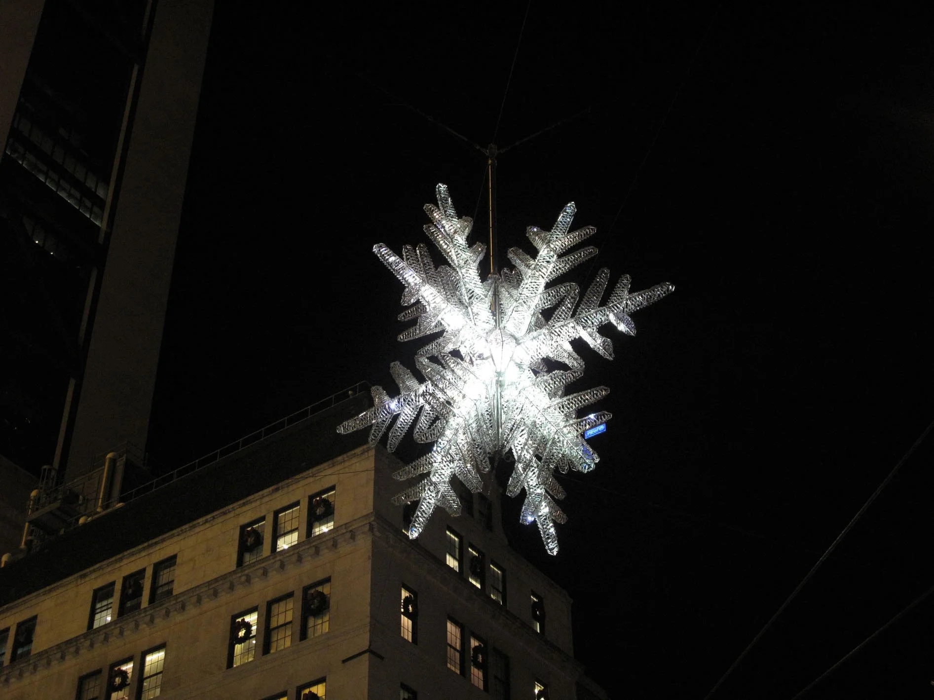 Illuminated snowflake-shaped holiday decoration hanging above a city building at night.