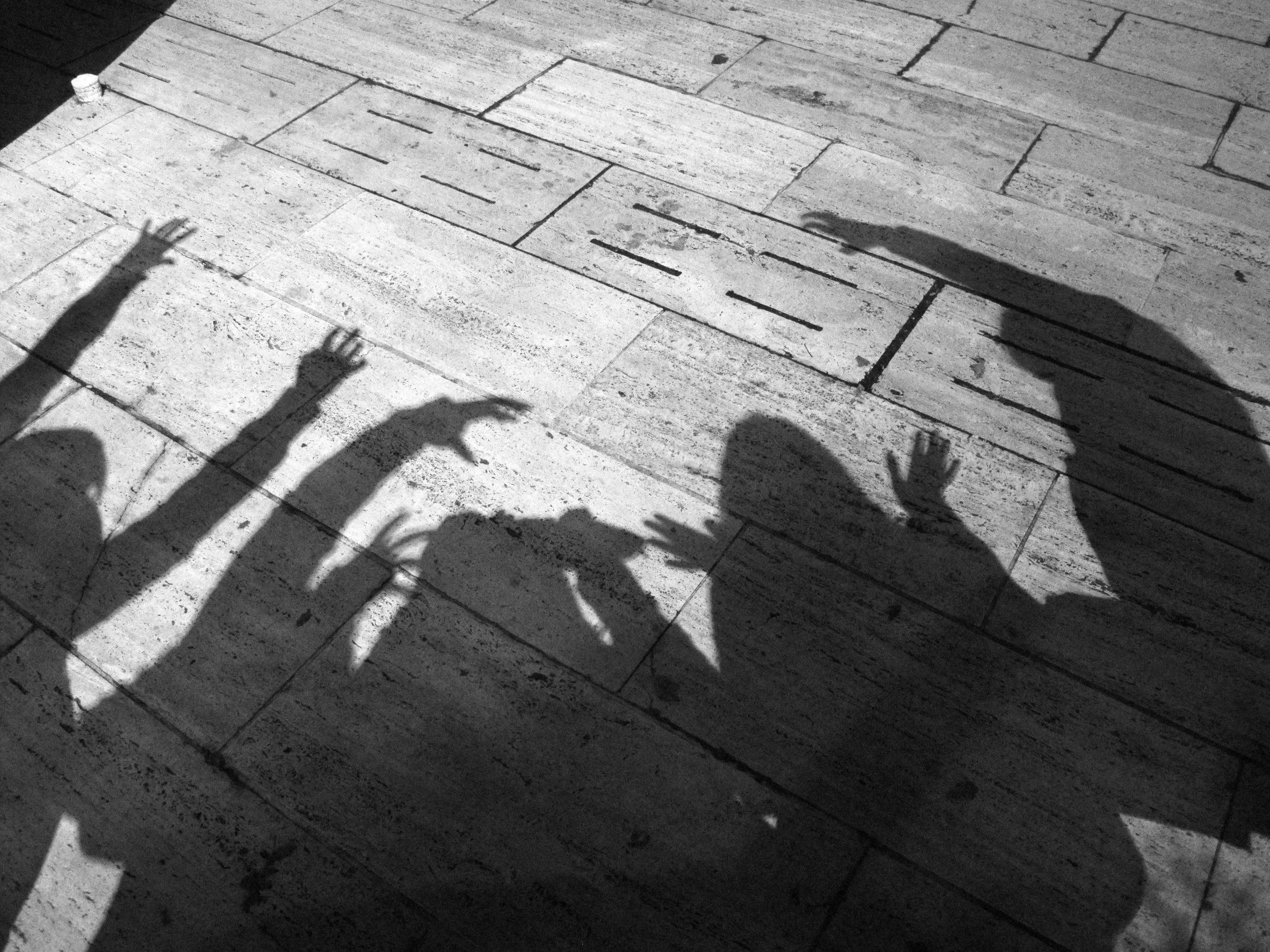 Shadows of three people on a stone pavement, with one person raising their hand and another making a gesture with their hand.