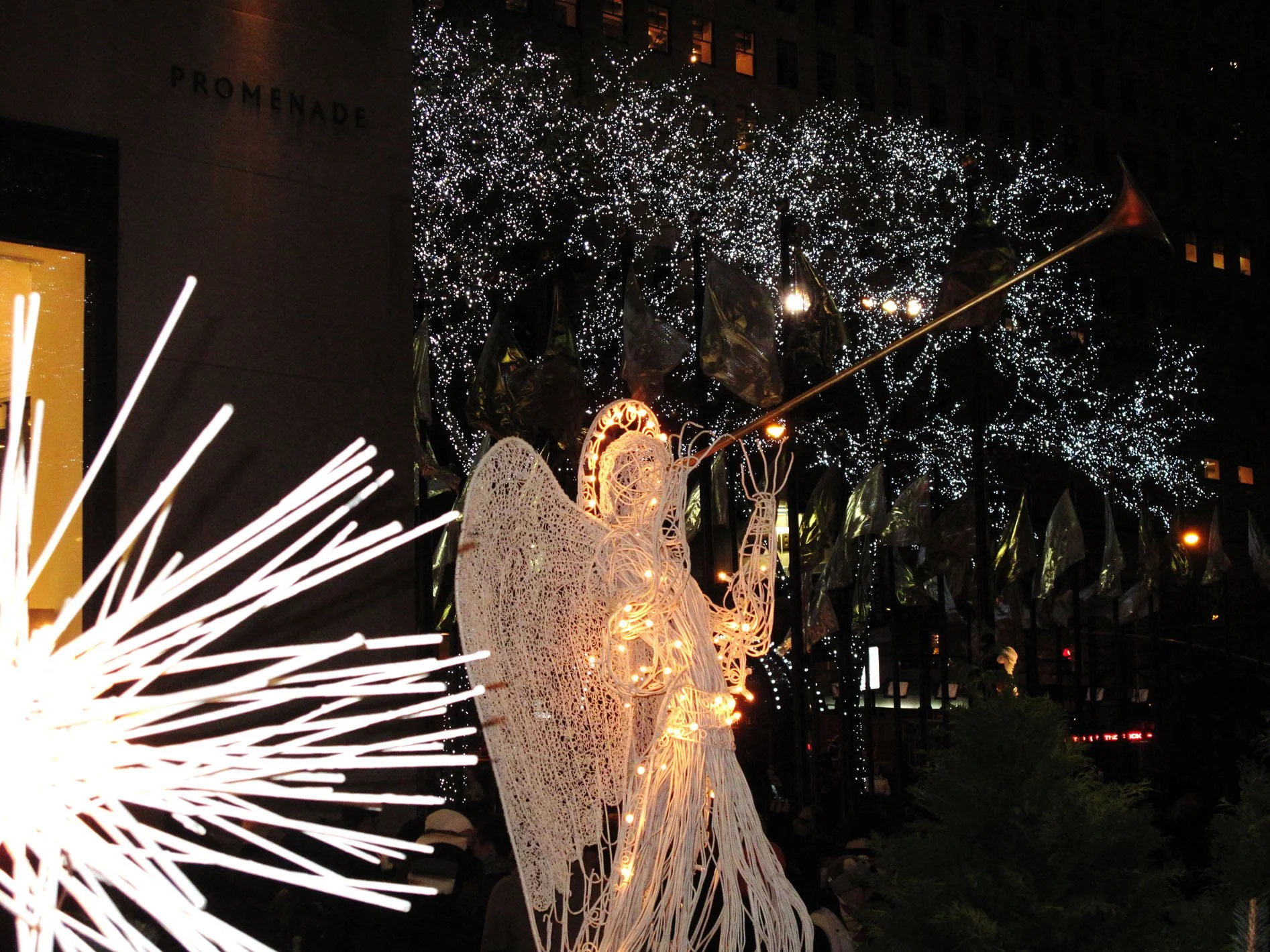 Nighttime holiday display featuring an illuminated angel figure with wings and a halo, holding a trumpet, surrounded by decorated trees with white lights and flags on poles.