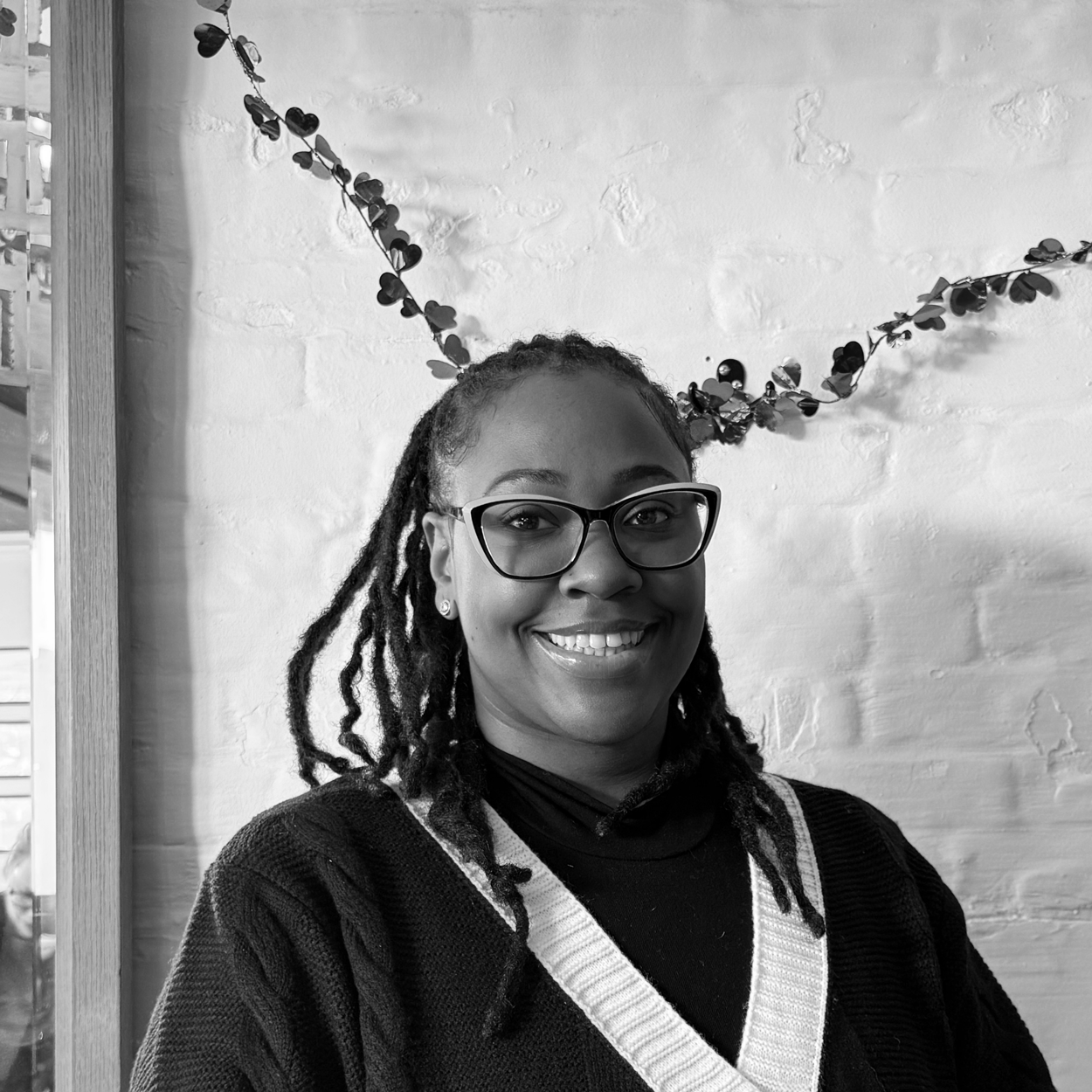 A smiling woman with glasses and dreadlocks wearing a dark sweater with a white V-neck and a black turtleneck underneath, sitting in front of a white brick wall decorated with a hanging garland of leaves.
